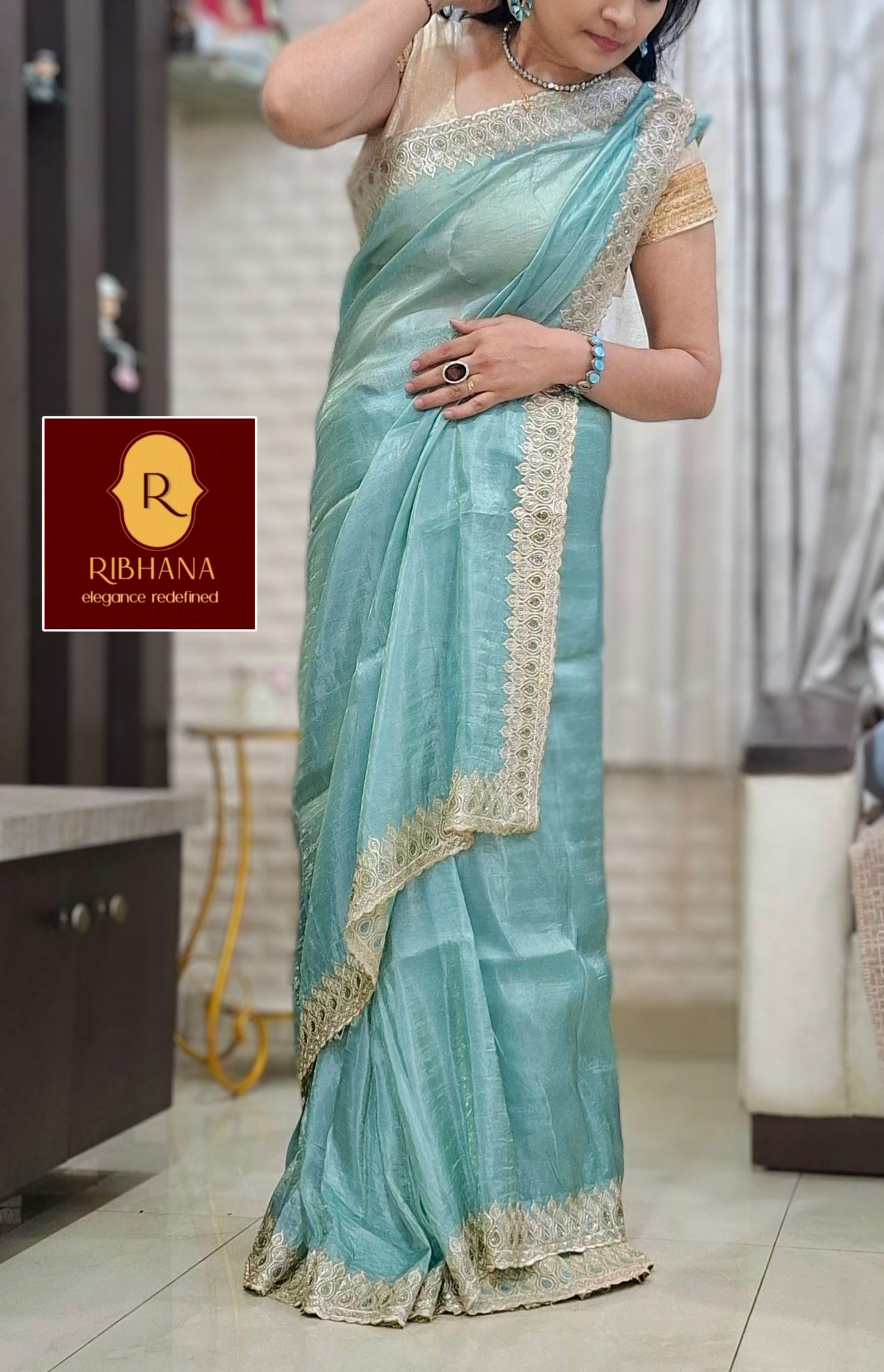A woman wearing a light blue-green silk saree with gold border embroidery, standing in an indoor setting. 