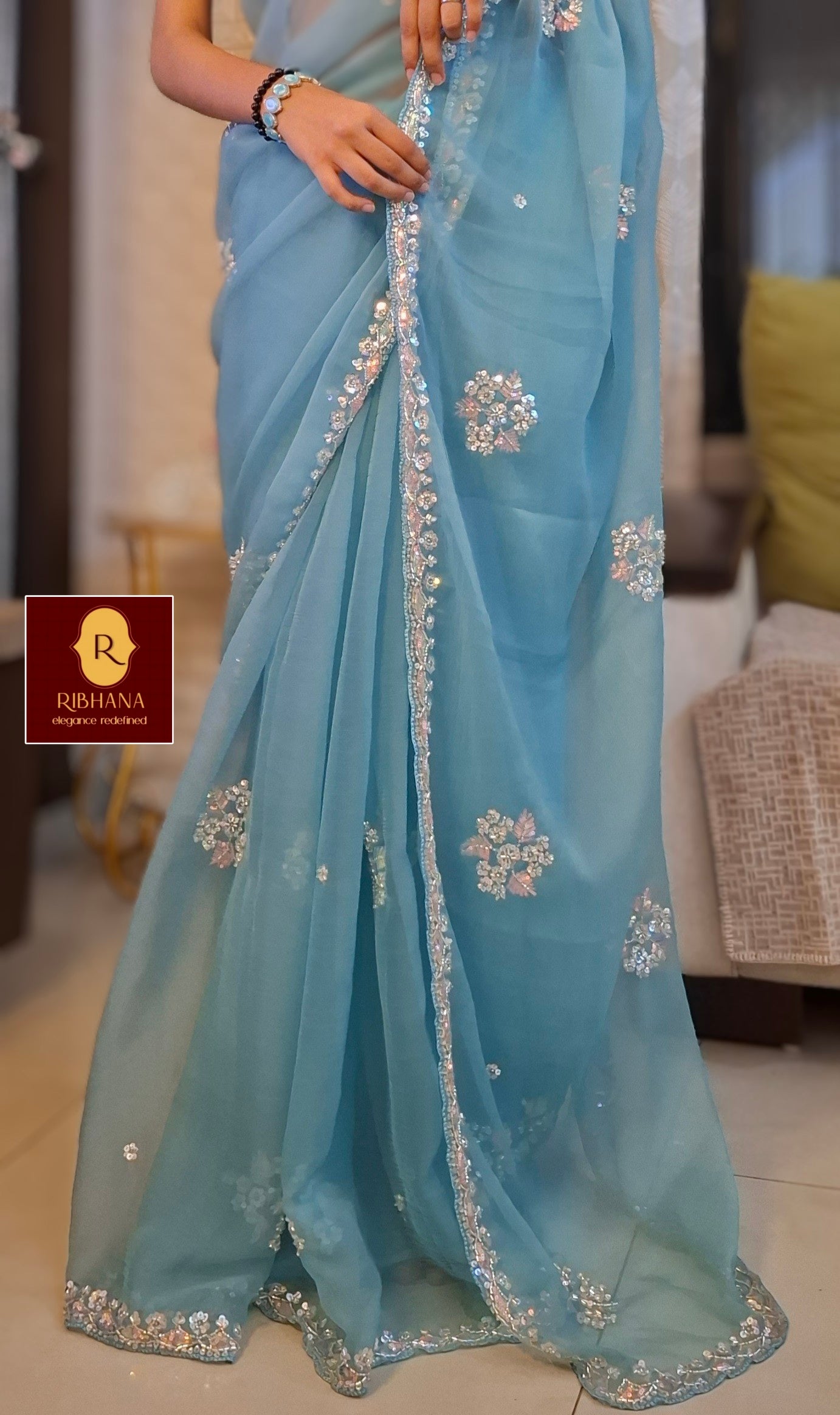 A light blue organza saree with intricate silver and white floral embroidery along the border and scattered motifs, worn by a model in an indoor setting. A brand logo for 'Ribhana' is visible in the bottom-left corner.