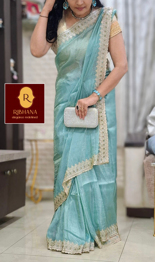A woman wearing a light blue-green silk saree with gold border detailing, standing in an indoor boutique setting. She is holding a small silver clutch purse.
