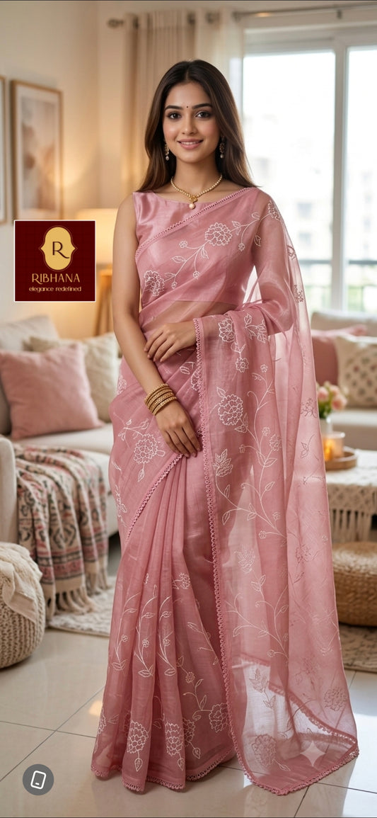A woman wearing a dusty pink embroidered saree with a matching sheer dupatta, standing in a well-lit living room. The brand logo 'Ribhana' is visible in the top left corner.