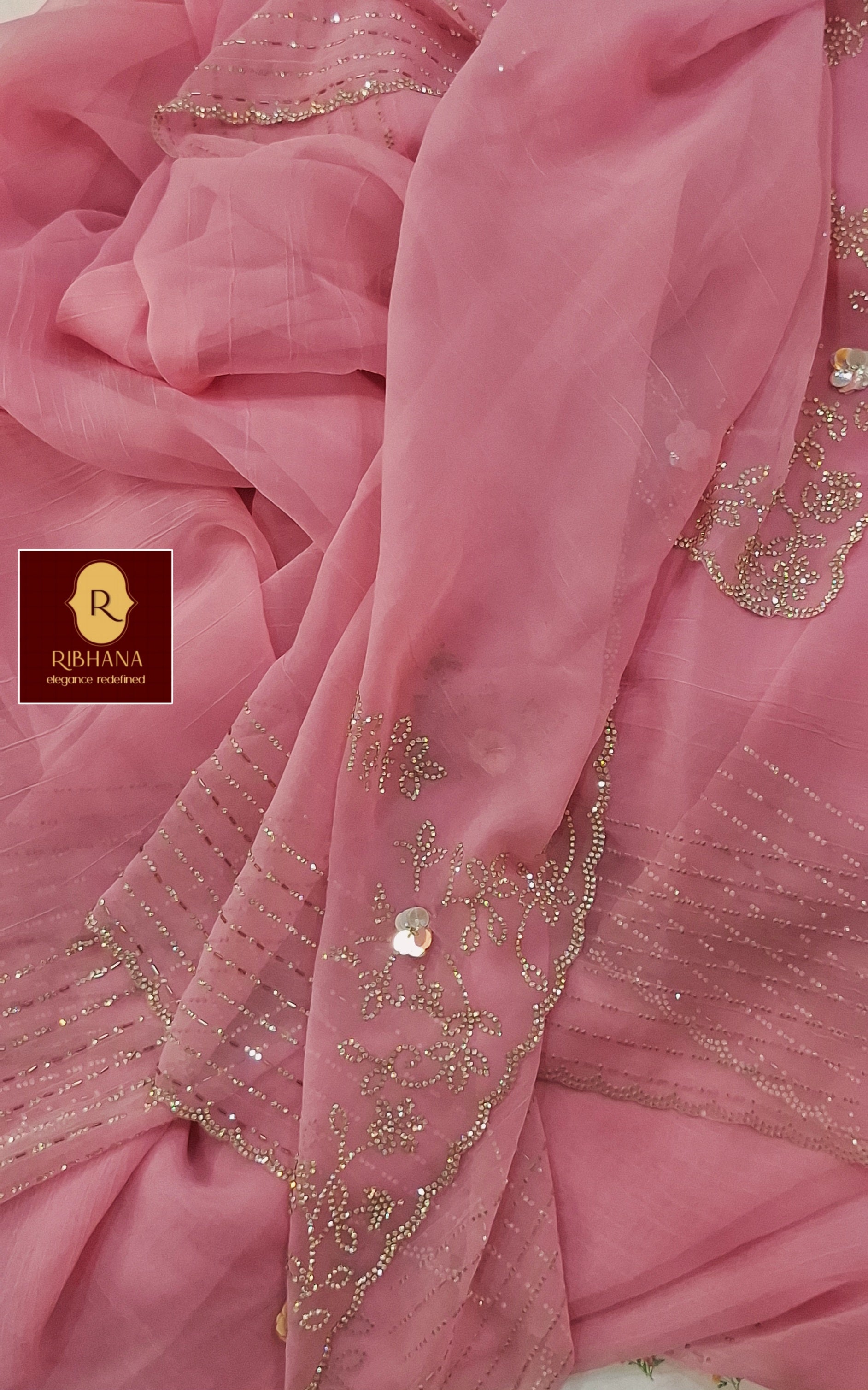 Pink organza fabric with gold sequin embroidery and mirror work, draped to show texture and border details. A logo for 'RIBHANA' is visible in the lower-left corner.