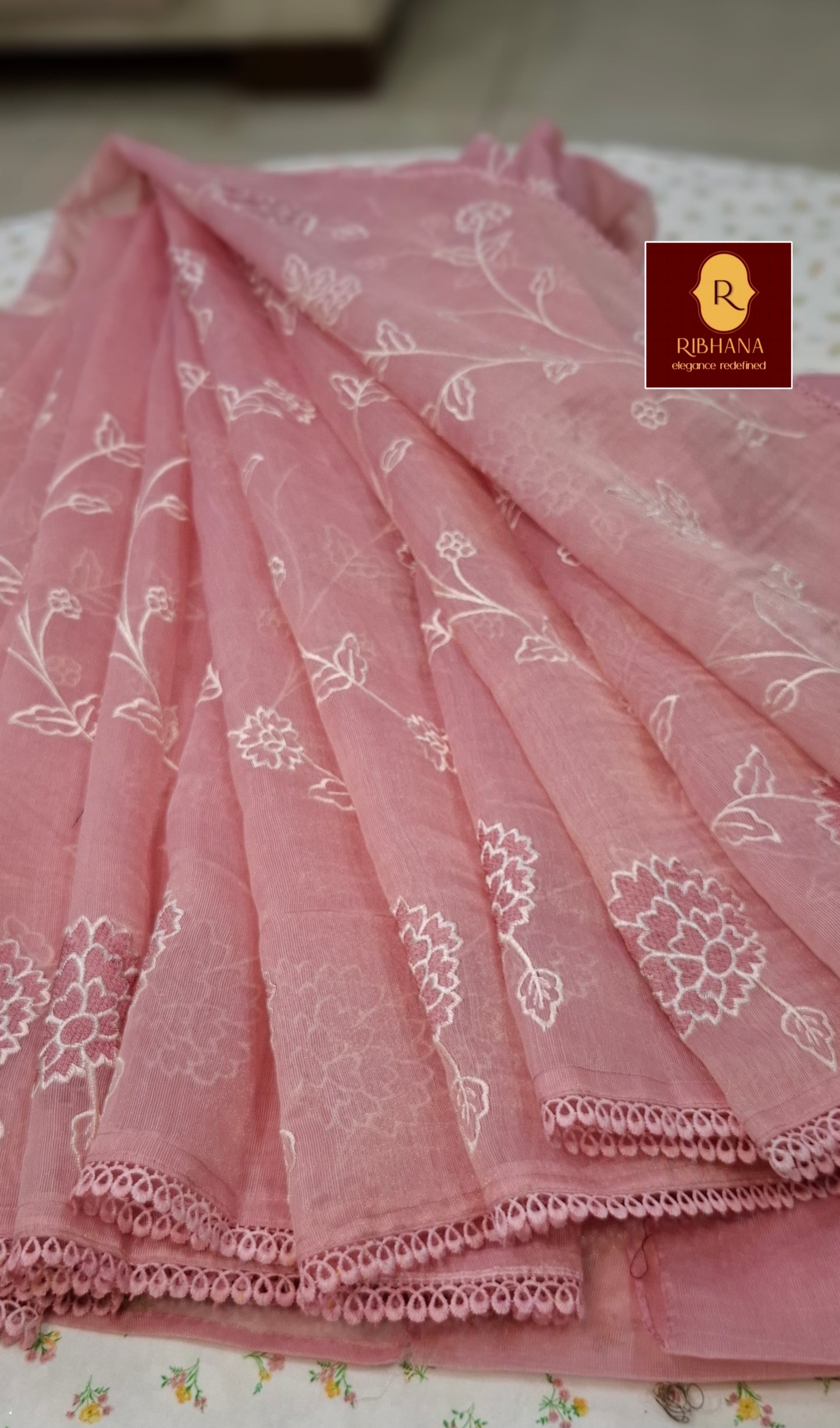 A pink sheer organza dupatta with white floral embroidery and a scalloped lace border, displayed in a draped arrangement. A logo for 'Ribhana' is visible in the top right corner.