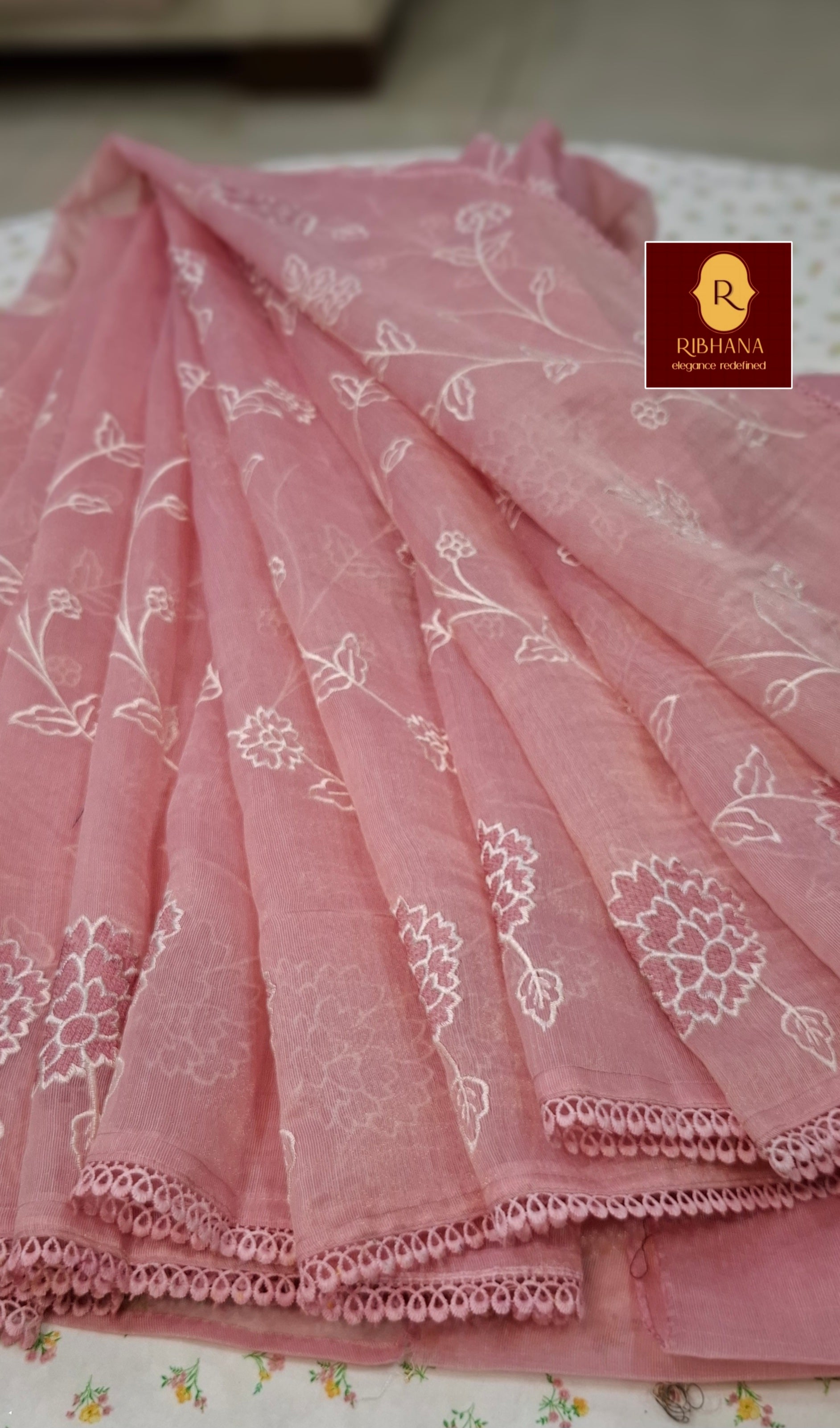 A pink sheer organza dupatta with white floral embroidery and a scalloped lace border, displayed in a draped arrangement. A logo for 'Ribhana' is visible in the top right corner.